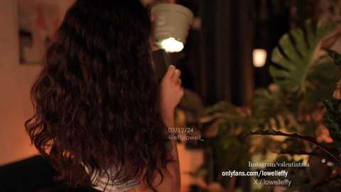 Media: Video of a woman with long, wavy brown hair, wearing a light-colored top, seen from behind, holding a drink in a dimly lit room with green plants in the background.