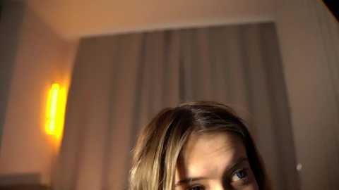 Media: Video of a young woman with light brown hair, partially visible, in a dimly-lit room with a warm, soft glow. The background features a large window with light gray curtains and a modern, vertical, orange lamp on the left.