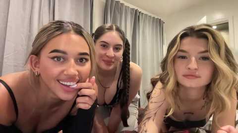 Media: Video of three young women with varying skin tones and hair colors, smiling, in a modern, softly lit bedroom.