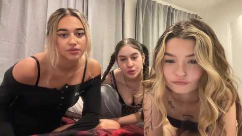 Media: Video of three young women with light skin and long, wavy hair, one blonde, one brunette, and one with blonde streaks, wearing casual, off-the-shoulder tops, leaning on a bed with red and white plaid sheets, in a dimly lit room with gray curtains.