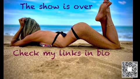 Media: Video of a tan, blonde woman in a black bikini, lying face-down on a sandy beach, legs bent, feet raised, blue sky background, playful text: \"The show is over. Check my links in bio.\