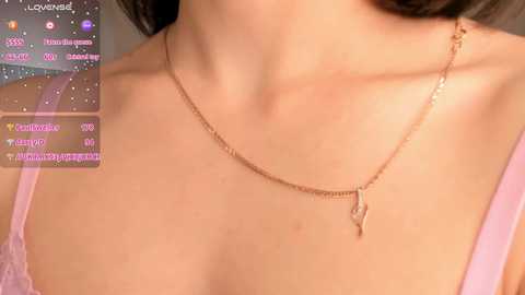 Media: A close-up video of a woman's bare shoulders and neck, adorned with a delicate gold necklace featuring a small, dangling heart pendant. Her skin is light, and she wears a soft, pink camisole. A live-streaming overlay with chat messages is visible in the top left corner.