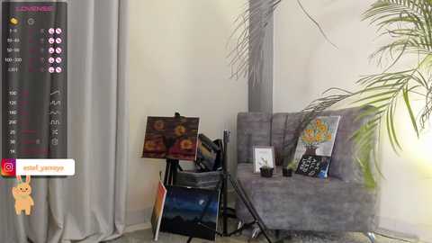 Media: A video of a cozy, modern living room with a gray sofa, a tall potted plant, an easel with an unfinished painting, and a small side table, all set against off-white walls.