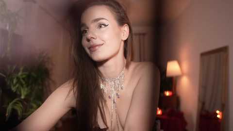 Media: Video of a smiling, topless woman with long brown hair, wearing a statement necklace, in a dimly-lit room with a potted plant and lit candles in the background.