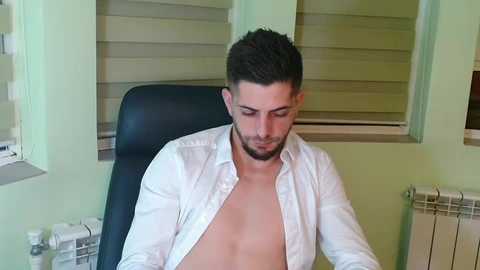 Media: Video of a shirtless, light-skinned man with short dark hair, wearing an open white button-up shirt, sitting in a black office chair, with beige window blinds and green walls in the background.