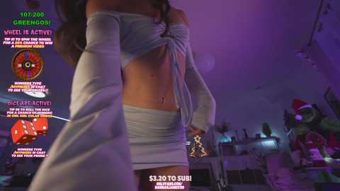 Media: Video of a slim woman with long brown hair in a tight white crop top and short skirt, taken indoors in a dimly lit room. Background includes gaming equipment and a Christmas tree.