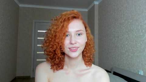 Media: Video of a fair-skinned woman with voluminous, curly, bright red hair, smiling, standing topless in a dimly lit room with beige wallpaper, a closed door, and a white bed visible in the background.