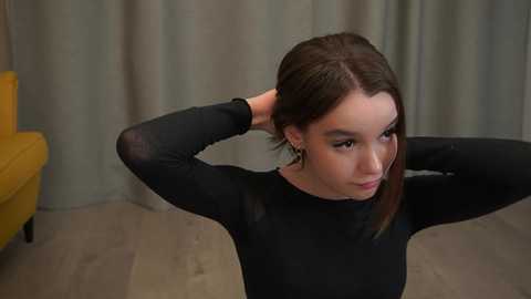 Video of a young woman with light skin, brown hair, and large hoop earrings, wearing a black long-sleeve top, adjusting her hair in a modern, casual indoor setting with a mustard-yellow couch and beige curtains.