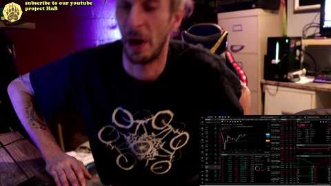 Media: Video of a man with a beard, wearing a black t-shirt with a white drone graphic, working on a computer in a dimly lit room with a brick wall, desk cluttered with cables, and a drone on a table.