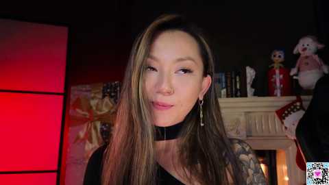 Media: Video of an Asian woman with long brown hair, wearing a black choker and tattoos, standing in a dimly lit room with festive decorations and a red light.