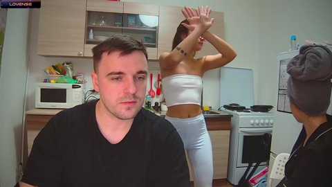 Media: Video of a man in a black shirt sitting on a couch, a woman in a white tube top and leggings dancing in a kitchen with wooden cabinets and a stove.