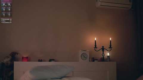 A video of a dimly-lit, minimalist bedroom with a white bed, a white dresser, and a black candelabra with lit candles. The background shows a beige wall and an air conditioning unit.