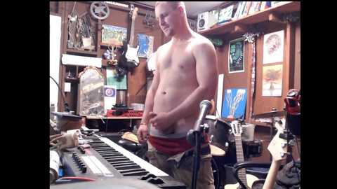 Media: A video of a shirtless, muscular man with a small penis, standing in a cluttered, dimly-lit room with a keyboard, posters, and various objects.