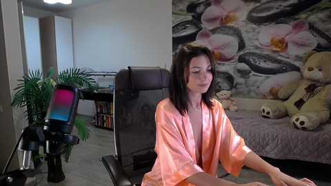 Media: Video of a young woman with shoulder-length dark hair, wearing a pink robe, sitting in a black office chair in a cozy room with a floral wall mural and plush teddy bear.