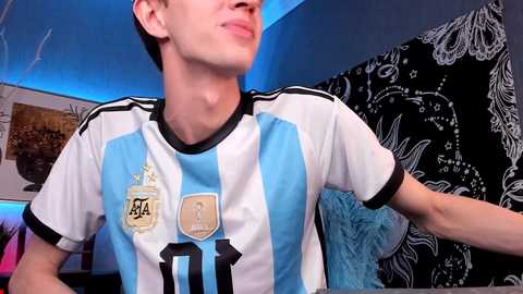 Media: Video of a young, fair-skinned, and thin man with short dark hair wearing a white and light blue Argentina soccer jersey, standing in a room with blue lighting, black and white wall art, and a blue blanket.