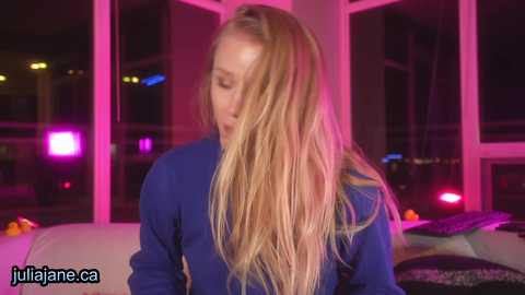 Media: Video of a blonde woman in a blue sweater, with long hair, indoors under pink and purple lighting, looking down, with blurred background objects.