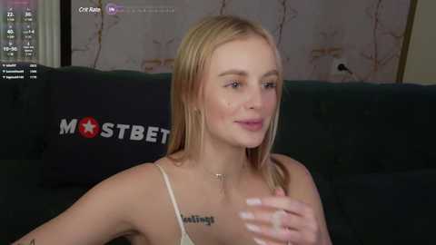 Media: Video of a blonde woman with a slender build and fair skin, wearing a white spaghetti-strap top, seated on a dark green couch. The background shows a muted floral wallpaper and a \"MOSTBET\" pillow. She has a serene expression.