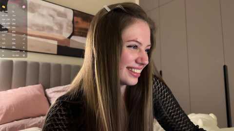 Media: Video of a smiling young woman with straight, light brown hair in a gray sweater, sitting on a bed with pink sheets and a beige headboard.