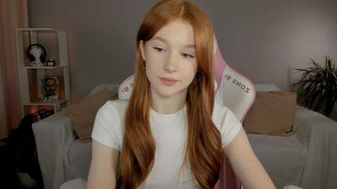 Media: Video of a young Caucasian woman with long, straight auburn hair, fair skin, and a slim physique, wearing a white T-shirt, sitting in a pink gaming chair in a dimly lit room with beige walls and a shelf holding books and a potted plant.