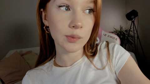 A close-up video of a young woman with fair skin, long red hair, and light blue eyes, wearing a white ribbed tank top. She has a subtle, natural makeup look. Background includes a bed with beige and brown pillows, a potted plant, and a black tripod.