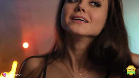 Media: Video of a young woman with long brown hair, light skin, and a slight smile, wearing a black top, set against a blurred, warm-toned background.