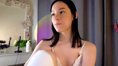 Media: A video of a young woman with straight, dark hair, wearing a white lace bra, standing in a brightly lit room with a mirror, fairy lights, and a colorful disco ball in the background.