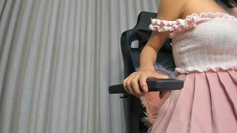 Media: Video of a woman with medium skin tone wearing a pink ruffled top and pleated skirt, gripping a black gaming chair, against a white curtain backdrop.