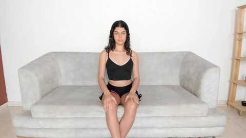 Media: Video of a young woman with long black hair, wearing a black sports bra and shorts, sitting on a light gray couch against a white wall in a simple room.