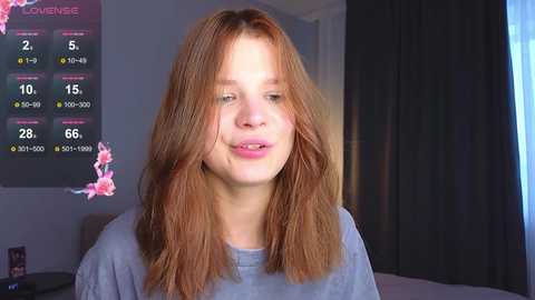 Media: Video of a young woman with light skin and shoulder-length auburn hair, smiling, wearing a gray t-shirt, in a dimly lit room with a TV displaying weather and cherry blossom decorations.