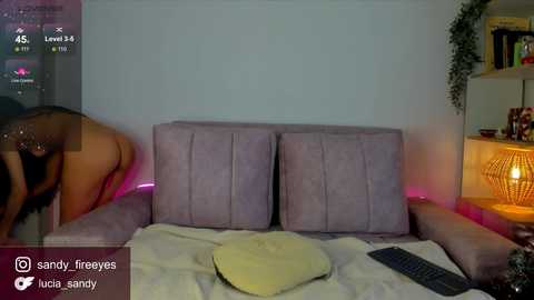 Media: Video of a woman in a sheer top bending over a pink couch, with a TV screen displaying a game, and a candlelit room in the background.