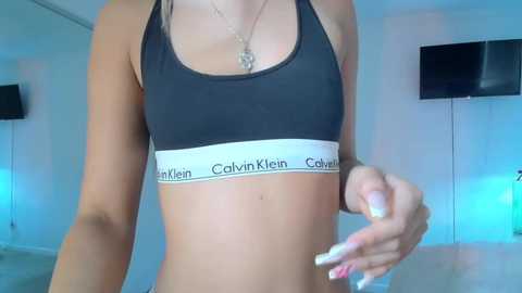 Media: Video of a slim, light-skinned woman in a black Calvin Klein sports bra, holding a bottle in a modern, minimally decorated room with two flat-screen TVs on white walls.