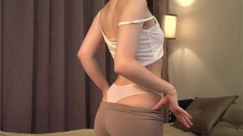 Media: Video of a slender, light-skinned woman in a white crop top and beige pants, pulling them down to reveal her thong, standing in a dimly lit room with beige walls and a lamp.