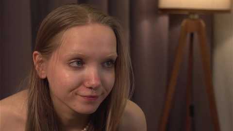 Media: Video of a young, fair-skinned, blonde-haired woman with a slight smile, wearing a light top, sitting indoors against dark curtains. A wooden tripod lamp is visible in the background.