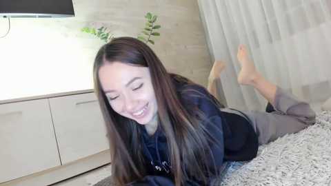 Media: A video of an East Asian woman with long black hair, lying on her stomach on a soft, light-colored rug, wearing a black hoodie and grey pants. She's smiling, barefoot, in a modern, minimalistic room with light wood cabinets and sheer curtains.