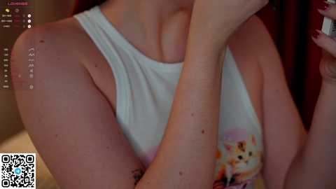 Media: Video of a fair-skinned woman with red hair, wearing a white tank top with a cartoon design, partially covering a tattoo on her arm.