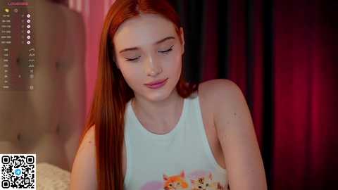 Media: Video of a fair-skinned, red-haired young woman with long hair, wearing a white tank top featuring a colorful graphic, sitting in a dimly lit room with a tufted headboard and dark curtains in the background.