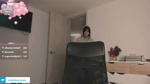 Media: Video of a young woman with long dark hair, wearing headphones, standing behind a black chair in a minimalist, modern living room with a flat-screen TV and a wooden side table.