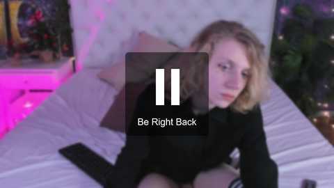 Media: Video of a blonde woman in black clothing, sitting on a bed with white sheets, watching a phone, with \"Be Right Back\" text overlay.