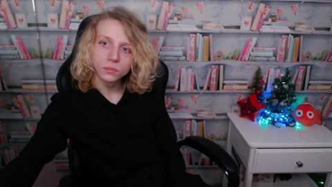 Media: Video of a blonde woman with curly hair in a black coat, seated in a chair, in a room with a bookshelf filled with books, and a small white table with blue and red ornaments and Christmas lights.