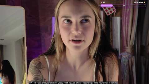 Media: Video of a young woman with fair skin and long, blonde hair, wearing a white tank top, standing indoors. She has a tattoo on her left arm and looks slightly anxious.