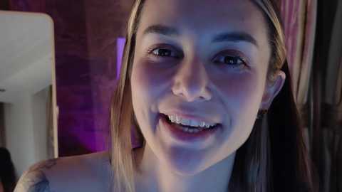 Media: Video of a smiling young woman with straight, light brown hair, wearing a white tank top, against a dimly lit, brick wall background.