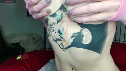 Media: Video of a person with fair skin, hands holding their chest revealing a colorful tattoo of a dragon and moon on the left side of their abdomen. Background includes a red blanket and a black cat.