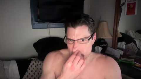 Media: A close-up video of a shirtless man with glasses, light skin, and short dark hair, covering his mouth in a bedroom setting.