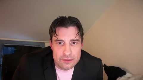 Media: Video of a light-skinned man with short, dark hair, wearing a black suit jacket over a light pink shirt, looking directly at the camera with a neutral expression. Background shows a beige wall and a partially visible black cat.