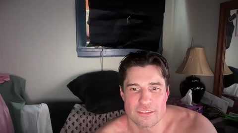 Media: Video of a shirtless man with short brown hair, smiling, in a dimly lit room with a TV and a lamp on a dresser.