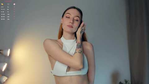 Media: Video of a slim, fair-skinned woman with long, straight brown hair, wearing a white halter top, smiling, with closed eyes, standing against a blurred background.