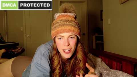 Media: Video of a smiling young woman with long brown hair, wearing a knit beanie and a blue shirt, lying on a bed in a dimly lit room.