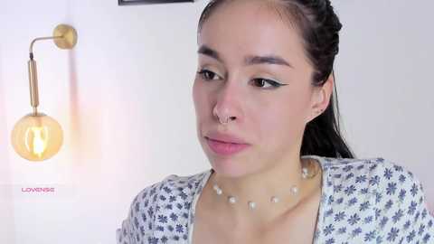 Media: Video of a young woman with light brown skin, braided hair, and dark eyes, wearing a white floral blouse, pearl necklace, and black eyeliner. Background features a wall lamp and white curtains.