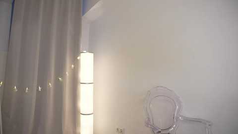 Media: A video of a minimalist bathroom with a white wall, a clear acrylic chair, a tall cylindrical lamp emitting a soft glow, and a white curtain partially drawn to the left.