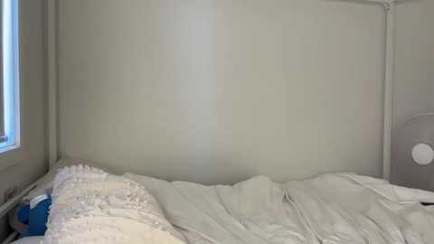 A video of a minimalist bedroom featuring a white wall with a faint shadow, a neatly made bed with white sheets, a small window, and a white fan on the right.